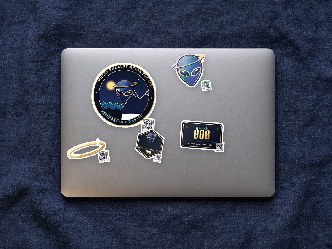 Saturday stickers on a laptop lid: the Surf | Sky badge, a holographic Alley, Drop 008, the gold ring and Saturday Original 001